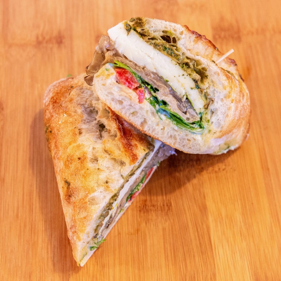 Close-up of a toasted ciabatta sandwich layered with roast beef, mozzarella, greens, tomato, and pesto on a wooden table.