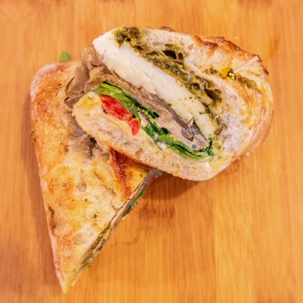 Open-faced sandwich on rustic bread with sliced meat, melted cheese, greens, tomato, and herb sauce on a wooden table.