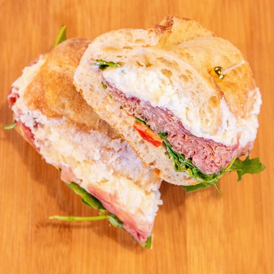 Two stuffed sandwiches with pink meat, creamy filling, tomatoes, and leafy greens on a wooden surface.
