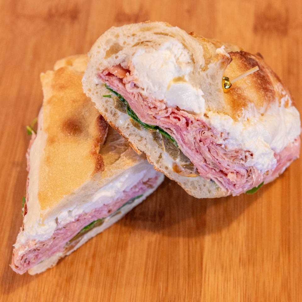 Sliced deli sandwich with pink meat, leafy greens, creamy filling, and toasted bread on a wooden table.