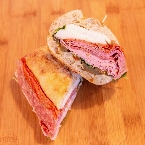 Two stacked deli sandwiches on bread, filled with layered pink and red meats, cheese, lettuce, and tomato on a wooden table.