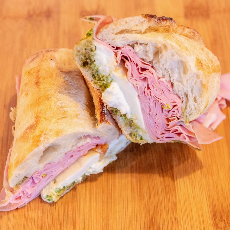 Sliced sandwich on rustic bread with pink noodles, creamy cheese, greens, and pickled onions on a wooden table.